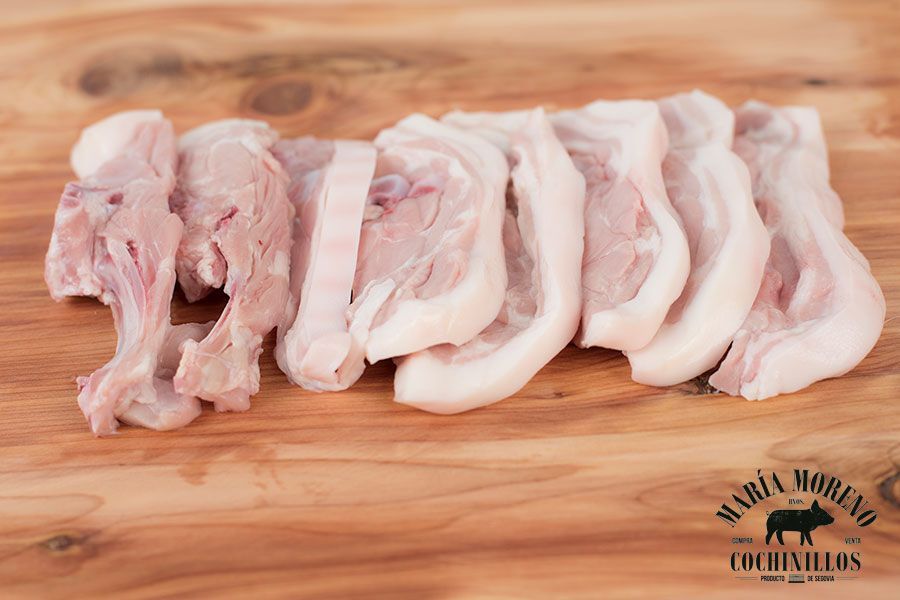 Las chuletillas de cochinillo de Segovia, unas tiernas costillas de cerdo con una capa de grasa, se presentan con gran arte sobre una tabla de cortar de madera. En la esquina inferior derecha, el logotipo de «María Moreno Cochinillos», acompañado de la silueta de un cerdo, destaca estos exquisitos cortes, ideales para cualquiera que desee comprar cochinillo de Segovia.
