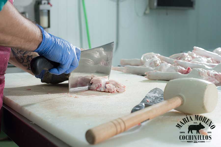 Una persona, que lleva guantes azules, utiliza un cuchillo de carnicero para cortar carne cruda sobre una mesa, con un mazo y varias porciones de carne a su lado: ideal para quienes quieran comprar cochinillo asado de Segovia. En la esquina inferior derecha se ve el logotipo de «María Moreno Cochinillos».