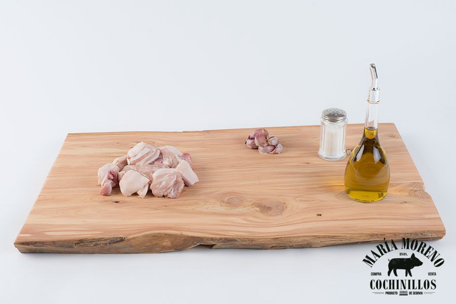 Sobre una tabla de cortar de madera, con un logotipo en la esquina inferior derecha, se apilan trozos de carne cruda, dientes de ajo, un salero y una botella de cristal de aceite de oliva, lo que evoca la tradición y la calidad que esperas cuando eliges el Cochifrito de cochinillo de Segovia.