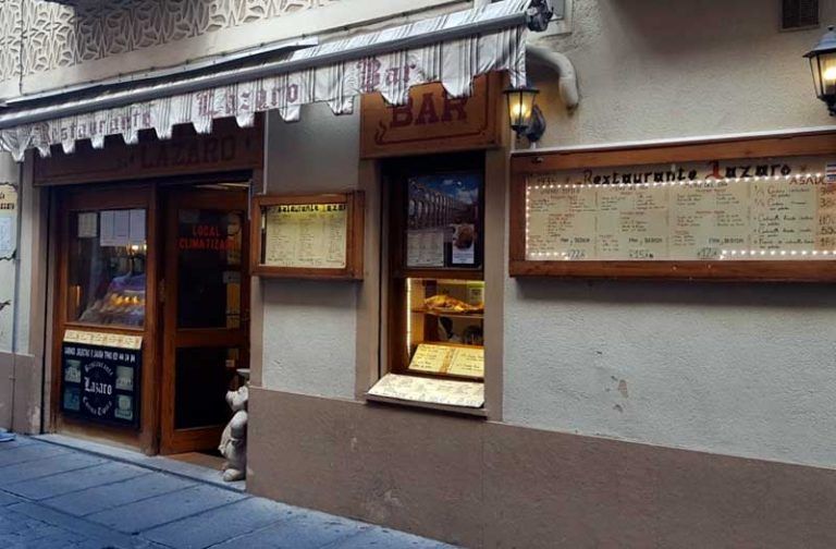 Exterior del Restaurante Lázaro, con una carta expuesta en la pared, una puerta marrón, una pequeña ventana y dos faroles. La entrada se encuentra en una calle estrecha: un lugar acogedor para quienes deseen comprar cochinillo de Segovia y disfrutar de la auténtica gastronomía local.