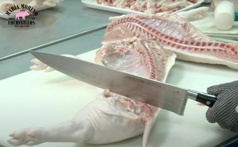 Un carnicero, que lleva un guante de malla metálica, utiliza un cuchillo grande para cortar una canal de cerdo cruda sobre una tabla de cortar blanca. En el fondo se ven otros trozos de carne, que recuerdan a la calidad que se encuentra al comprar cochinillo de Segovia.
