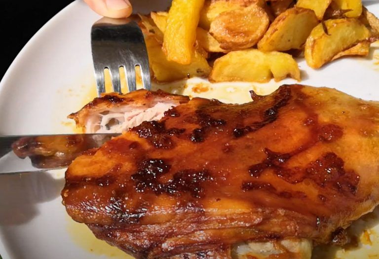 Una mano utiliza un tenedor y un cuchillo para cortar un trozo de pollo asado glaseado en un plato blanco, con patatas asadas doradas al fondo: una escena que recuerda a quienes buscan comprar cochinillo de Segovia para disfrutar de una experiencia gastronómica verdaderamente auténtica.