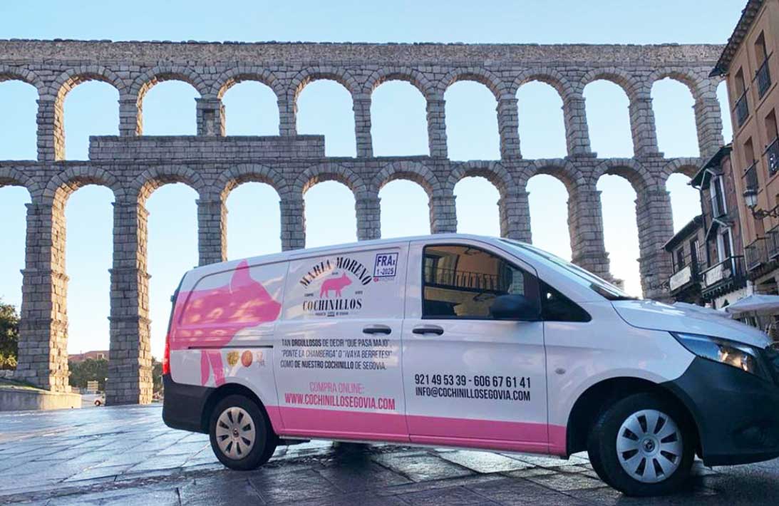 Una furgoneta de reparto blanca y rosa está aparcada frente al antiguo acueducto romano de Segovia, en España, en un día soleado; quizá lista para repartir delicias locales a quienes quieran comprar cochinillo de Segovia.